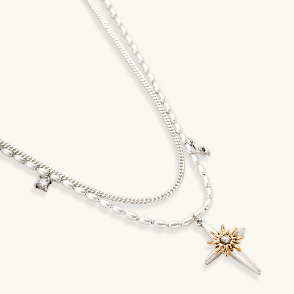Two-layer silver-toned necklace with star charms and dagger pendant featuring golden sun motif on white background.