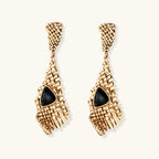 Gold-toned geometric mesh earrings with black triangular resin stones and fringe detail on neutral background