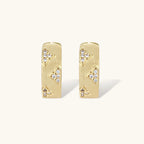 Gold-plated hoop earrings with textured surface and raised floral motifs set with clear zirconia stones on cream background.
