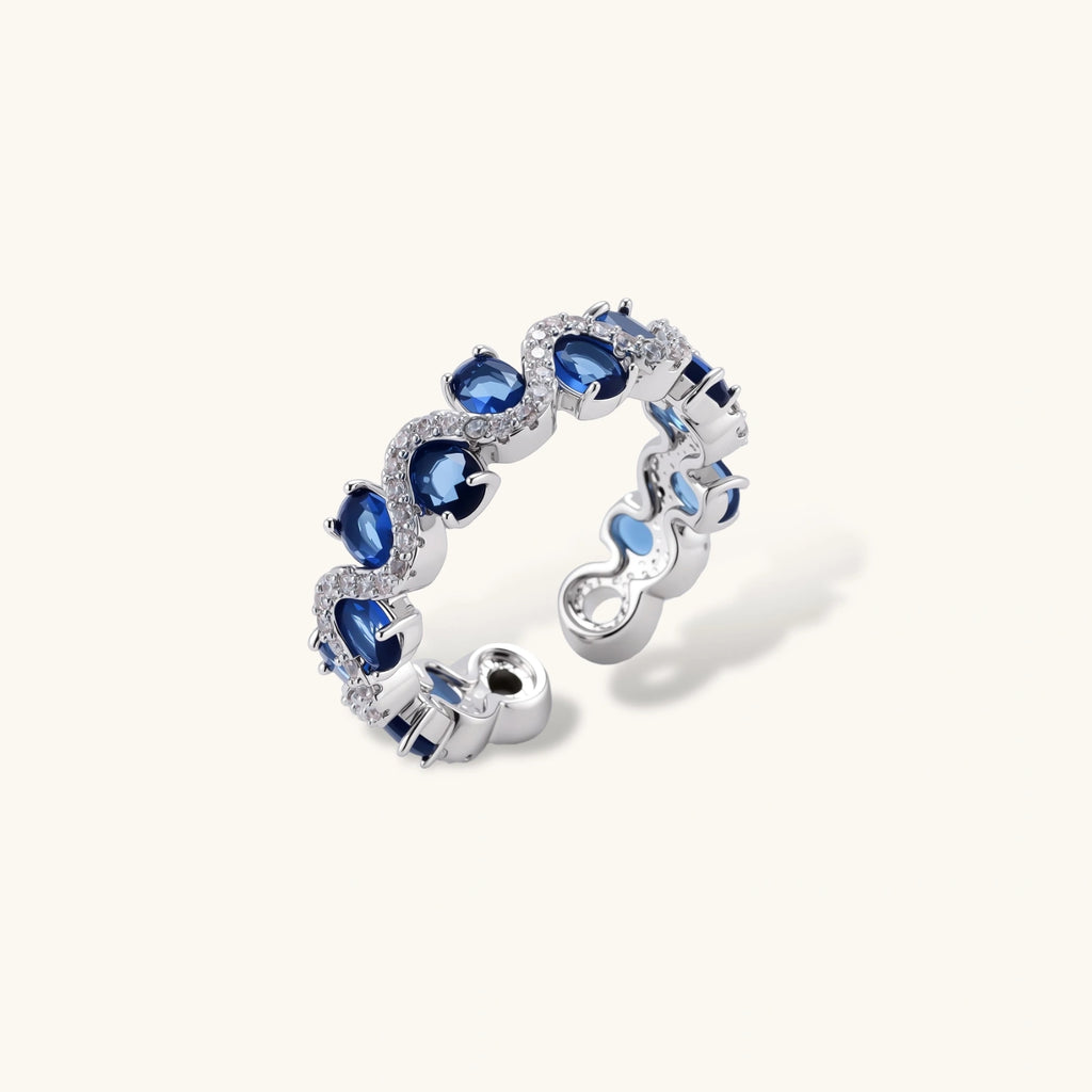 Adjustable ring with blue cubic zirconia stones in flowing wave pattern, copper with platinum plating on white background.
