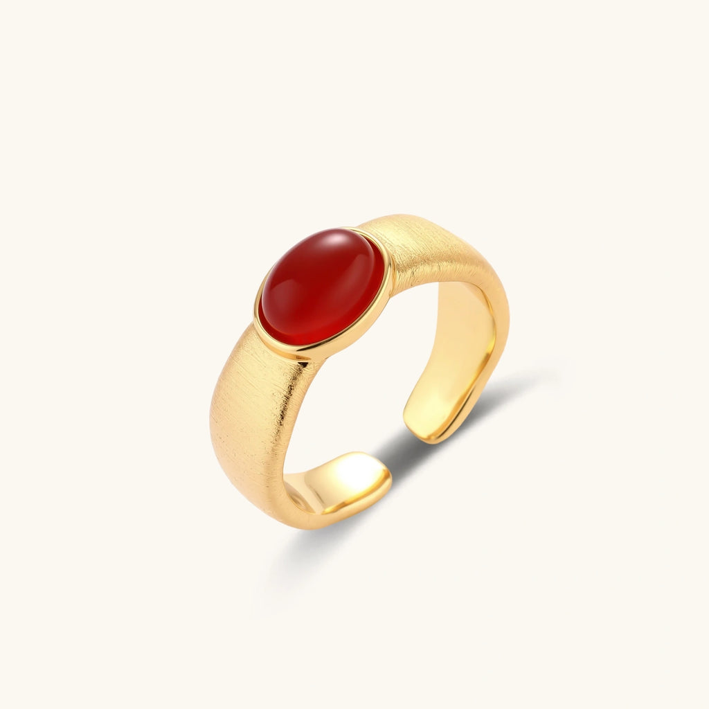 Close-up of brushed gold-plated ring with deep red oval stone, featuring adjustable band design on neutral background.